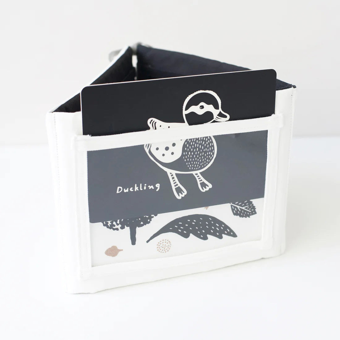 A TUMMY TIME GALLERY soft book from WEE GALLERY, featuring black and white fish and squid illustrations, is partially inserted in a clear front pocket. A brown tag attached by a string reads "tummy time gallery." This Montessori toy, ideal for sensory development, rests on a plain white surface.