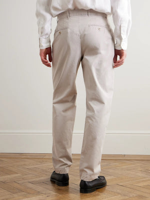 A person wearing HARTFORD - TAYLOR PANTS IN STONE, a white long-sleeve shirt, and black loafers stands on a wooden floor against a neutral wall.