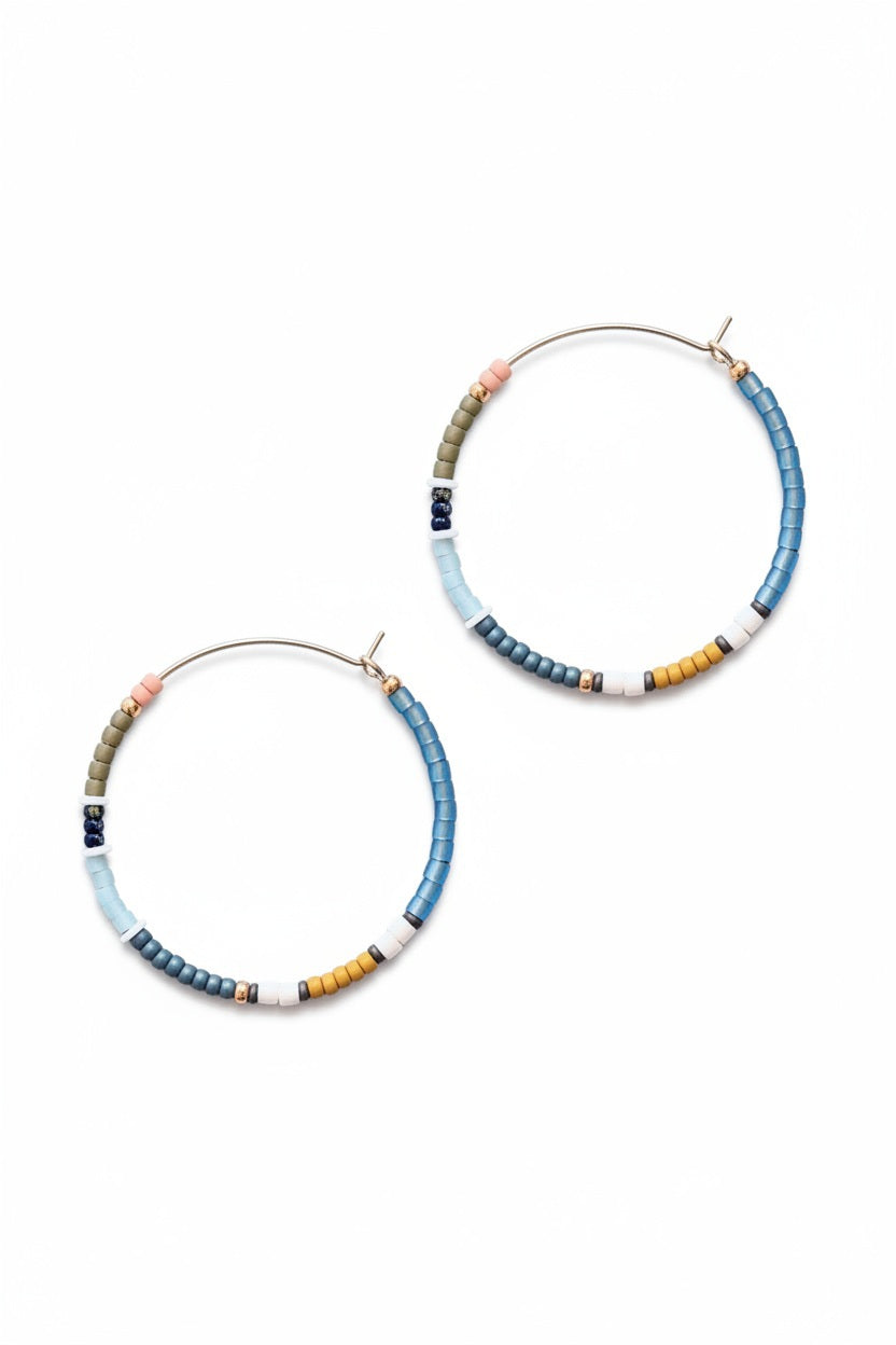 The ALICE RISE - BIG COLORLOOP EARRINGS IN SPRINGWATER by ALICE RISE feature two large hoops adorned with small, colorful Japanese glass beads, elegantly arranged and displayed on a textured light grey surface.