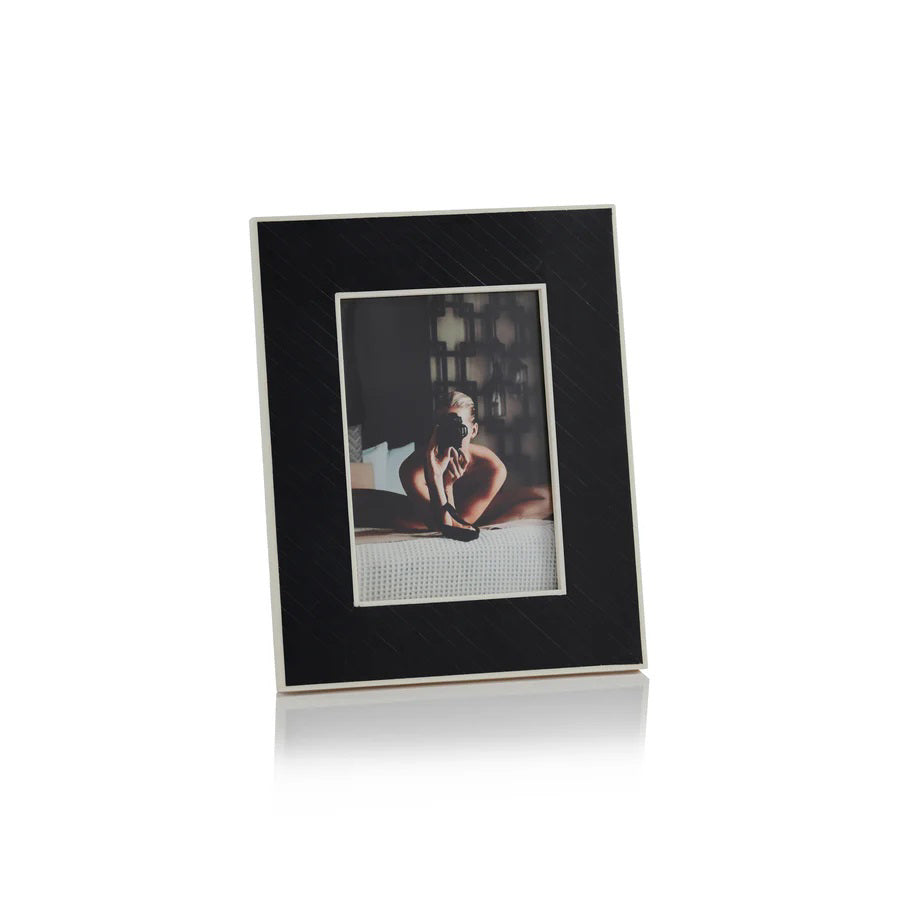 The ZODAX BLACK BONE INLAY WITH WHITE INNER BORDER is a black, rectangular 5x7 picture frame featuring a white inner border, shown upright against a white background displaying a photo of a person sitting on a bed.
