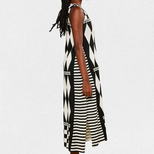 A person is wearing the IN BED WITH YOU - LONG KNIT DRESS featuring a sleeveless, knee-length design and a striking geometric pattern. The dress, from the Italian brand IN BED WITH YOU by CLAUDIOLAI SRL, showcases alternating squares and circles in vibrant prints of red, yellow, purple, and white set against a plain white textured background.