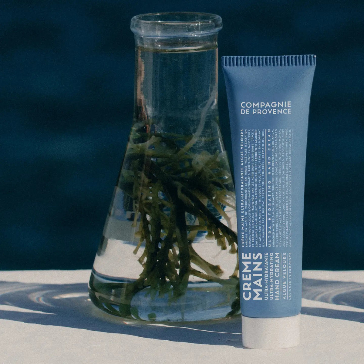 A blue tube of CIE LUXE BRANDS COMPAGNIE DE PROVENCE - TRAVEL HAND CREAM VELVET SEAWEED 10Z stands next to a beaker of water and green seaweed, set against a dark blue background to showcase its botanical oils for lasting hydration.
