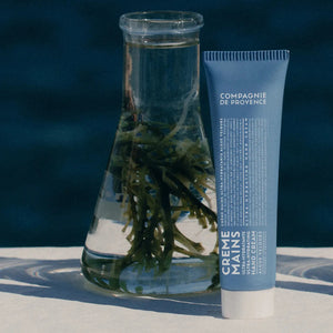 A blue tube of CIE LUXE BRANDS COMPAGNIE DE PROVENCE - TRAVEL HAND CREAM VELVET SEAWEED 10Z stands next to a beaker of water and green seaweed, set against a dark blue background to showcase its botanical oils for lasting hydration.