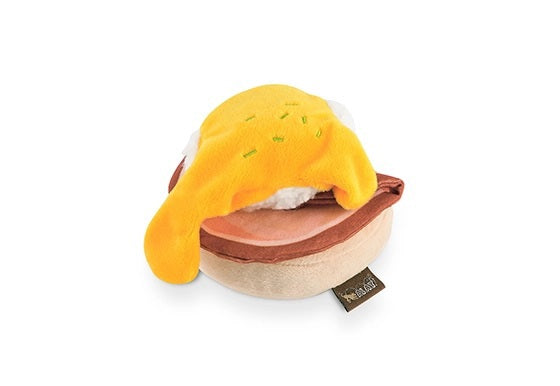 Introducing the PLAY PET LIFESTYLES/BRANDS BARKING BRUNCH - BENNY’S BENEDICT: an adorable dog toy designed to look like a plush egg with a yellow yolk atop a round, brown base, mimicking a classic brunch favorite. Perfect for playful pups, this fun and charming brunch-themed toy features a black fabric tag attached at the bottom.