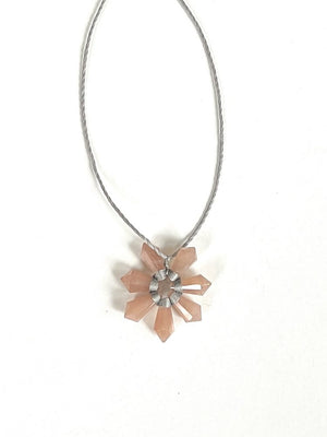 The ANN LIGHTFOOT - PEACH MOONSTONE SEVEN POINT FLOWER NECKLACE features a thin, gray braided cord and a pendant with light pink peach moonstone crystal points in a flower-like arrangement.