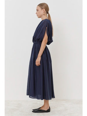 Wearing the ankle-length dark blue MEIMEIJ - OVERLAY DRESS, this person stands against a white backdrop. The dress features gathered sleeves and an elasticized waist, accessorized with black flats and large round earrings, while their hair is neatly pulled back.