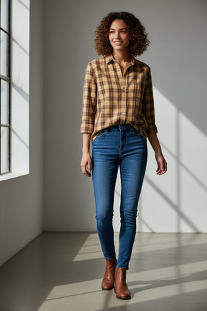 A woman stands in a sunlit room, smiling with curly hair, wearing the FRANK & EILEEN EILEEN Relaxed Button Up Shirt in Italian Flannel Gold Brown Plaid, paired with blue skinny jeans and brown ankle boots.