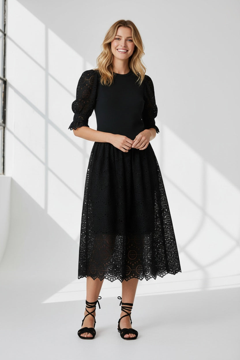 A woman stands against a white background wearing the SFIZIO - BRODERIE ANGLAISE MIDI DRESS by SFIZIO, featuring puffed sleeves and a lace skirt, paired with black strappy sandals.