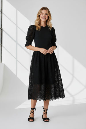 A woman stands against a white background wearing the SFIZIO - BRODERIE ANGLAISE MIDI DRESS by SFIZIO, featuring puffed sleeves and a lace skirt, paired with black strappy sandals.