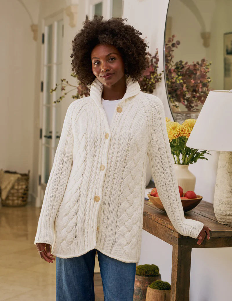 The FRANK &amp; EILEEN Cotswolds Cable Knit Cardigan in ivory is a chunky Italian cotton sweater, featuring a ribbed shawl collar, braided cable knit pattern, and brown buttons.