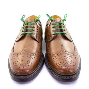 A pair of brown leather brogues with decorative perforations feature premium STOLEN RICHES Dress Shoelaces in Niklaus Green, displayed facing forward on a white background.
