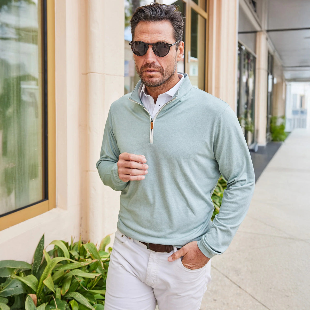 A man with short dark hair and a beard wears sunglasses and the COLLARS AND CO - PALM PIMA BLEND QUARTER ZIP IN MINT over a white collared shirt, leaning against a light-colored wall outdoors, showcasing modern comfort in this pullover.
