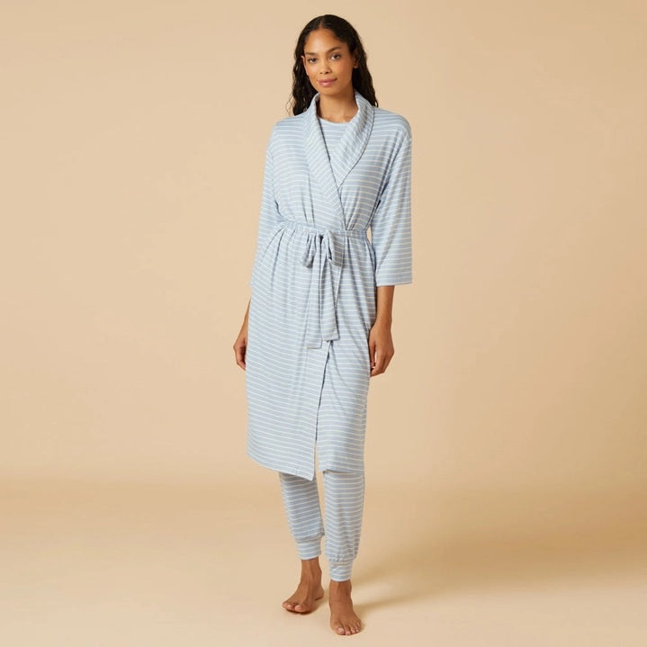 A woman wearing the SOFTIES Dream Shawl Collar Robe in Sky Blue Stripe by SOFTIES sits on a wooden chair, adjusting her hair with one hand and resting her other arm on the chair’s back against a beige background, evoking spa-like luxury akin to slipping into this lightweight robe.