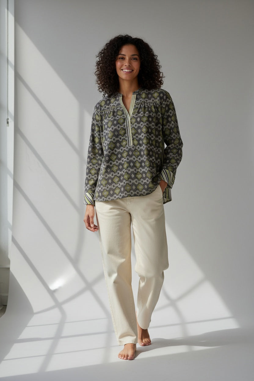 A woman with curly hair smiles while standing barefoot in sunlight, wearing the STORIATIPIC Margot Blouse in Anise—a patterned, long-sleeve cotton design from STORIATIPIC—paired with light-colored pants; perfect for a gentle machine wash at 30 degrees.