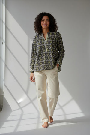 A woman with curly hair smiles while standing barefoot in sunlight, wearing the STORIATIPIC Margot Blouse in Anise—a patterned, long-sleeve cotton design from STORIATIPIC—paired with light-colored pants; perfect for a gentle machine wash at 30 degrees.