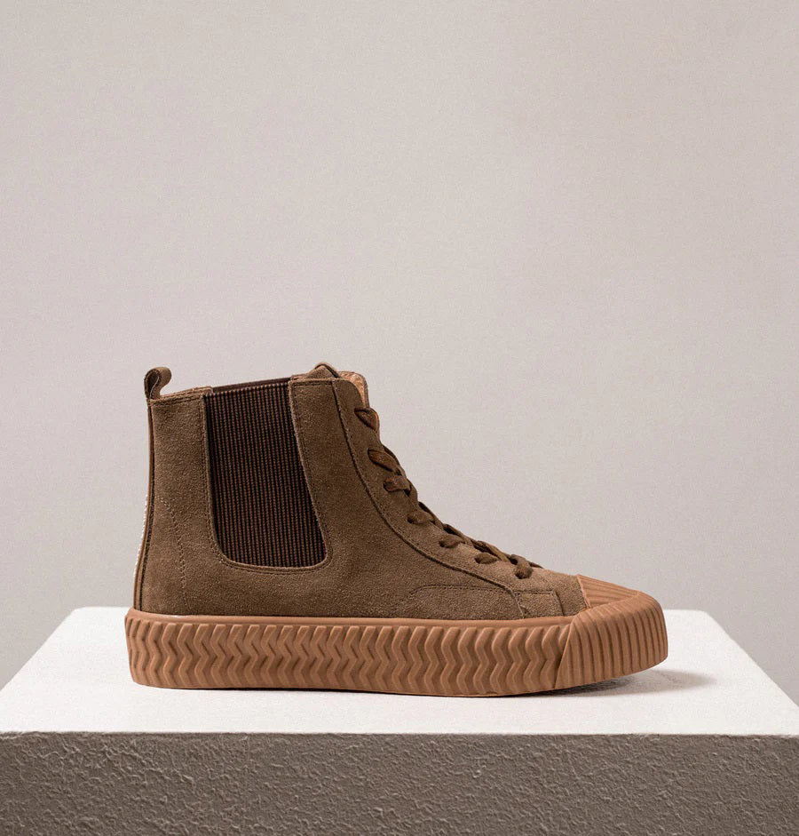 A pair of DANIELLA LEHAVI - OSAKA HIGH TOP SNEAKERS with a contemporary design, featuring brown suede uppers and rugged rubber soles, are placed side by side on a white pedestal against a plain beige background. These comfortable high-top sneakers boast a lace-up design and ribbed toe caps for added durability.