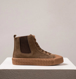 A pair of DANIELLA LEHAVI - OSAKA HIGH TOP SNEAKERS with a contemporary design, featuring brown suede uppers and rugged rubber soles, are placed side by side on a white pedestal against a plain beige background. These comfortable high-top sneakers boast a lace-up design and ribbed toe caps for added durability.