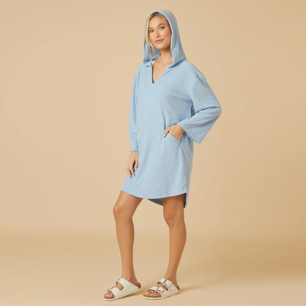 A woman faces away in the SOFTIES Terry Hooded Lounger in Heather Sky Blue, paired with white sandals. Her blonde ponytail grazes a visible bikini top, blending resort-ready comfort and style against a beige backdrop.