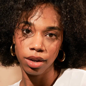 A close-up of a person with natural curly hair, adorned in gold hoop earrings and a white top. They wear a neutral facial expression while looking directly at the camera. The softly lit, plain background enhances their glowing skin as if touched by JASON WU - TINTED MOISTURIZER MEETS CC CREAM from JASON WU BEAUTY.