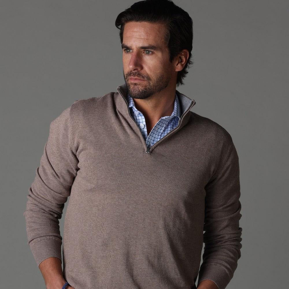A man with dark hair and a beard wears the COLLARS AND CO - SEMI SPREAD COLLAR POLO IN BLUE TATTERSAL under a light brown sweater, standing against a gray background and looking to his right with a serious expression.
