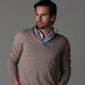 A man with dark hair and a beard wears the COLLARS AND CO - SEMI SPREAD COLLAR POLO IN BLUE TATTERSAL under a light brown sweater, standing against a gray background and looking to his right with a serious expression.