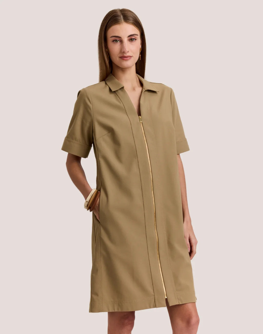 A woman wears the HINSON WU BRIDGET Short Sleeve Dress in Traveler’s Twill Khaki, featuring a collar and zip front. She pairs it with gold hoop earrings and bangles, standing against a plain light background.