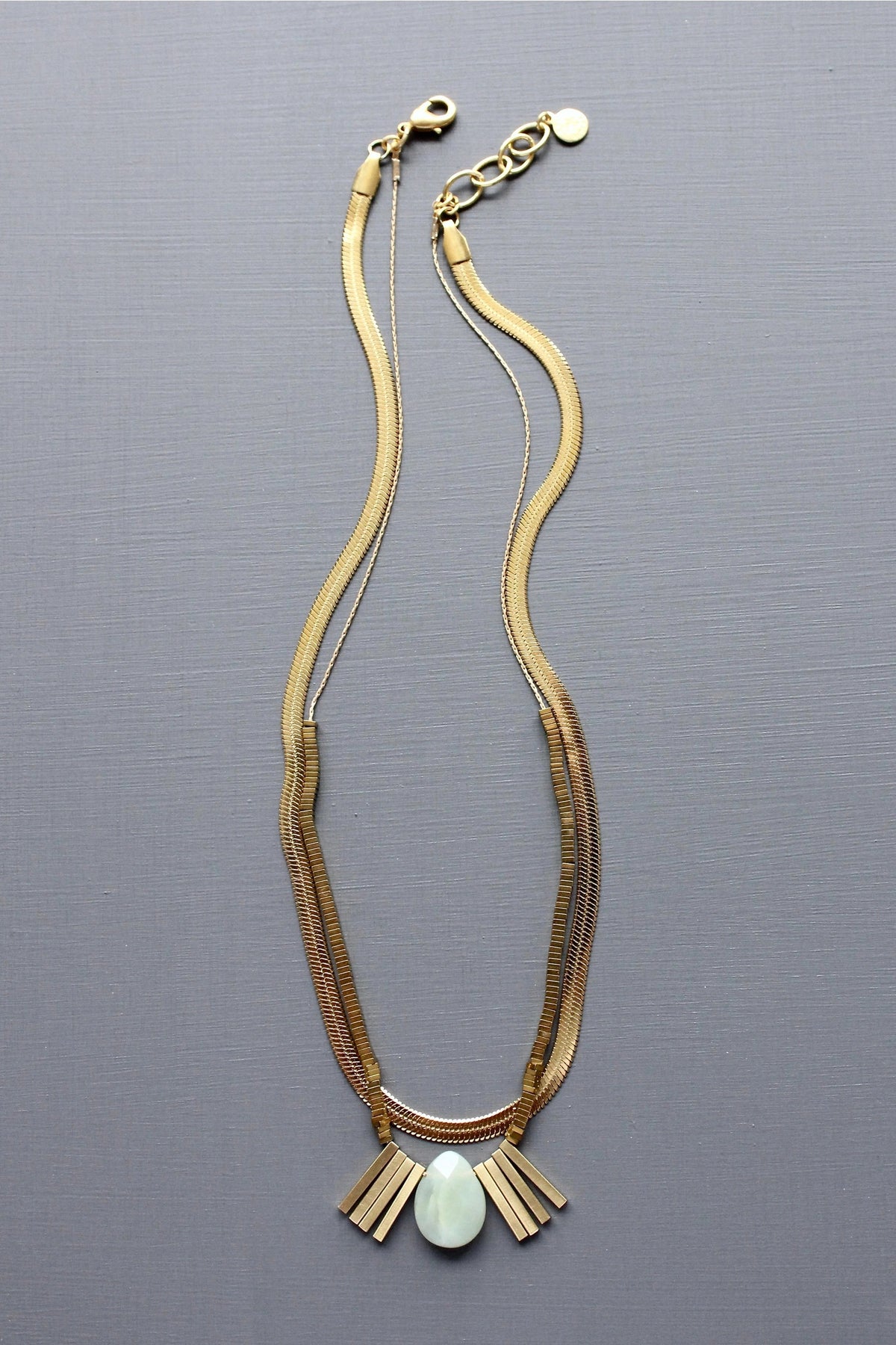 The DAVID AUBREY DOUBLE STRAND SNAKE CHAIN AND JADE ARTDECO PENDANT NECKLACE features two gold-tone layered chains, a large oval jade pendant, and four small gold rectangular accents, beautifully displayed on a gray background.
