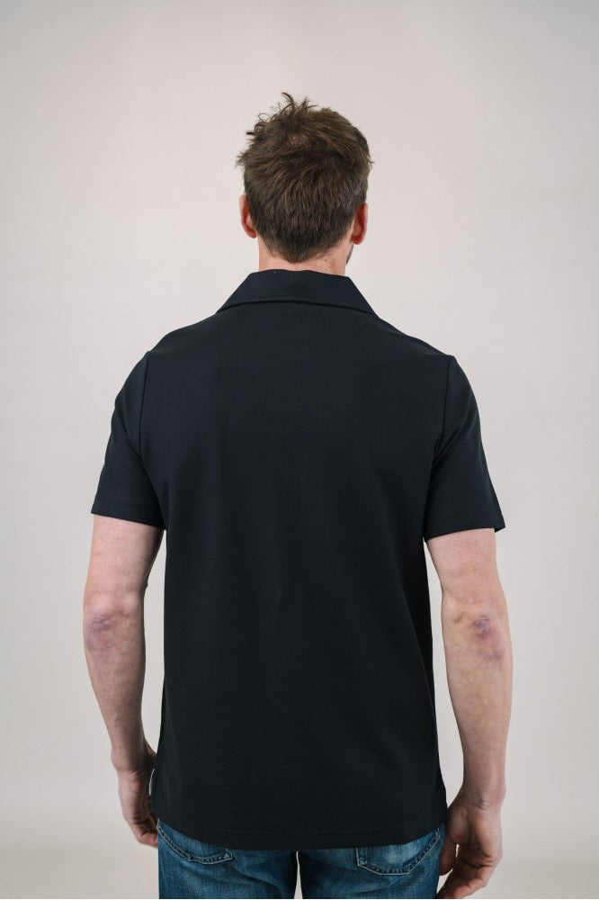 A man with short brown hair and a trimmed beard models the EDWARD ARMAH Pima Cotton Tailored Polo Shirt in white with light-colored pants, standing against a plain light background, one hand in his pocket, looking to the side.