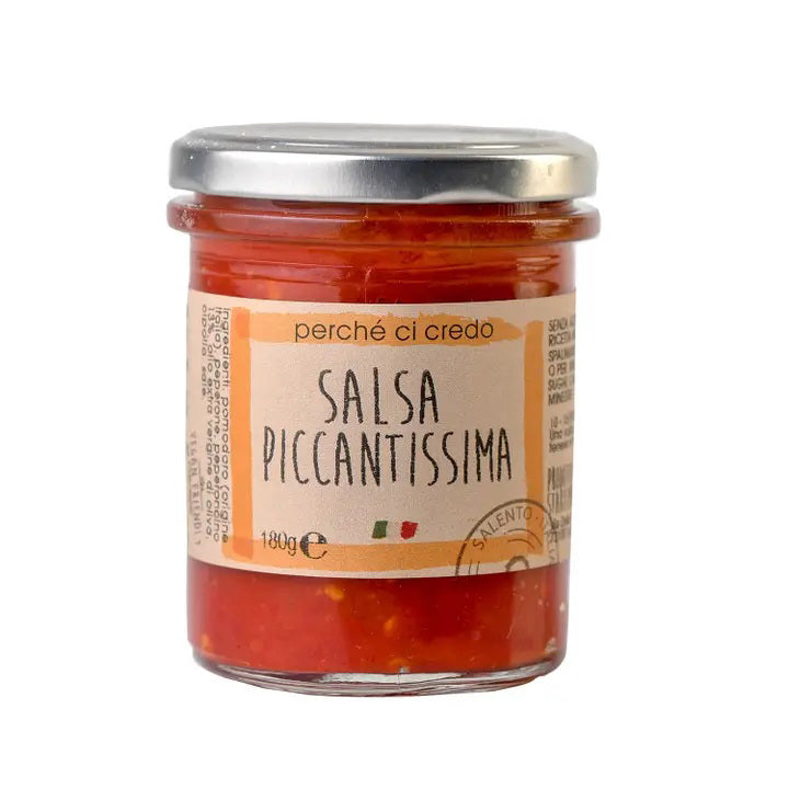 A small glass jar of PERCHE CI CREDO - SALSA PICCANTISSIMA EXTRA SPICY SPREAD from ZIA PIA IMPORTS, featuring a metallic lid. The jar contains 180 grams of bright red, chunky sauce made from fresh ingredients. The beige label with orange borders showcases text along with small Italian flag icons.