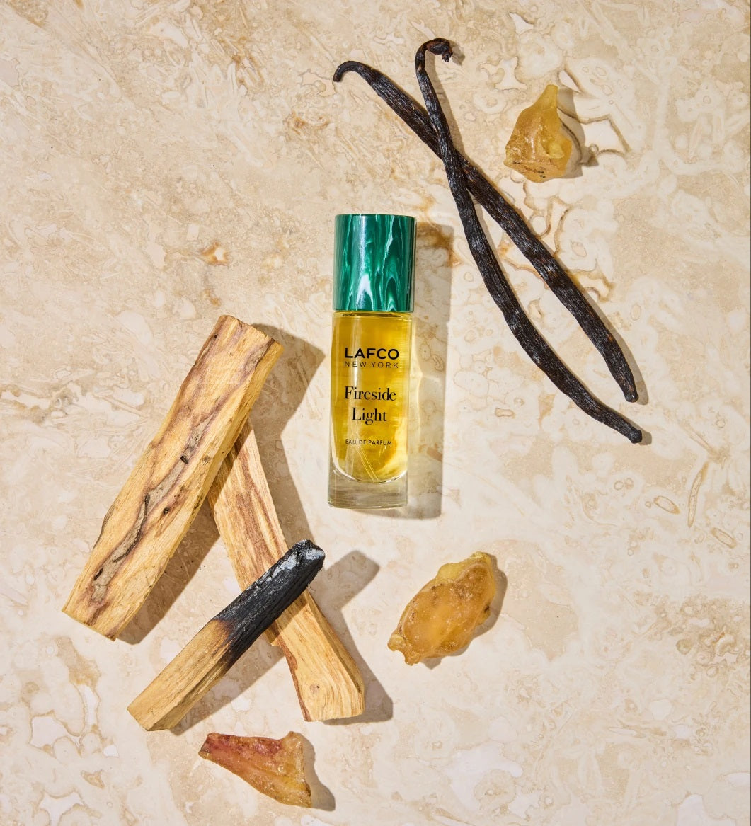 A 10ml bottle of LAFCO Fine Fragrance, Fireside Light rests atop a tree stump beside a vanilla bean, its green cap and brown smoky background evoking the warm woody notes of this vegan and cruelty-free scent.