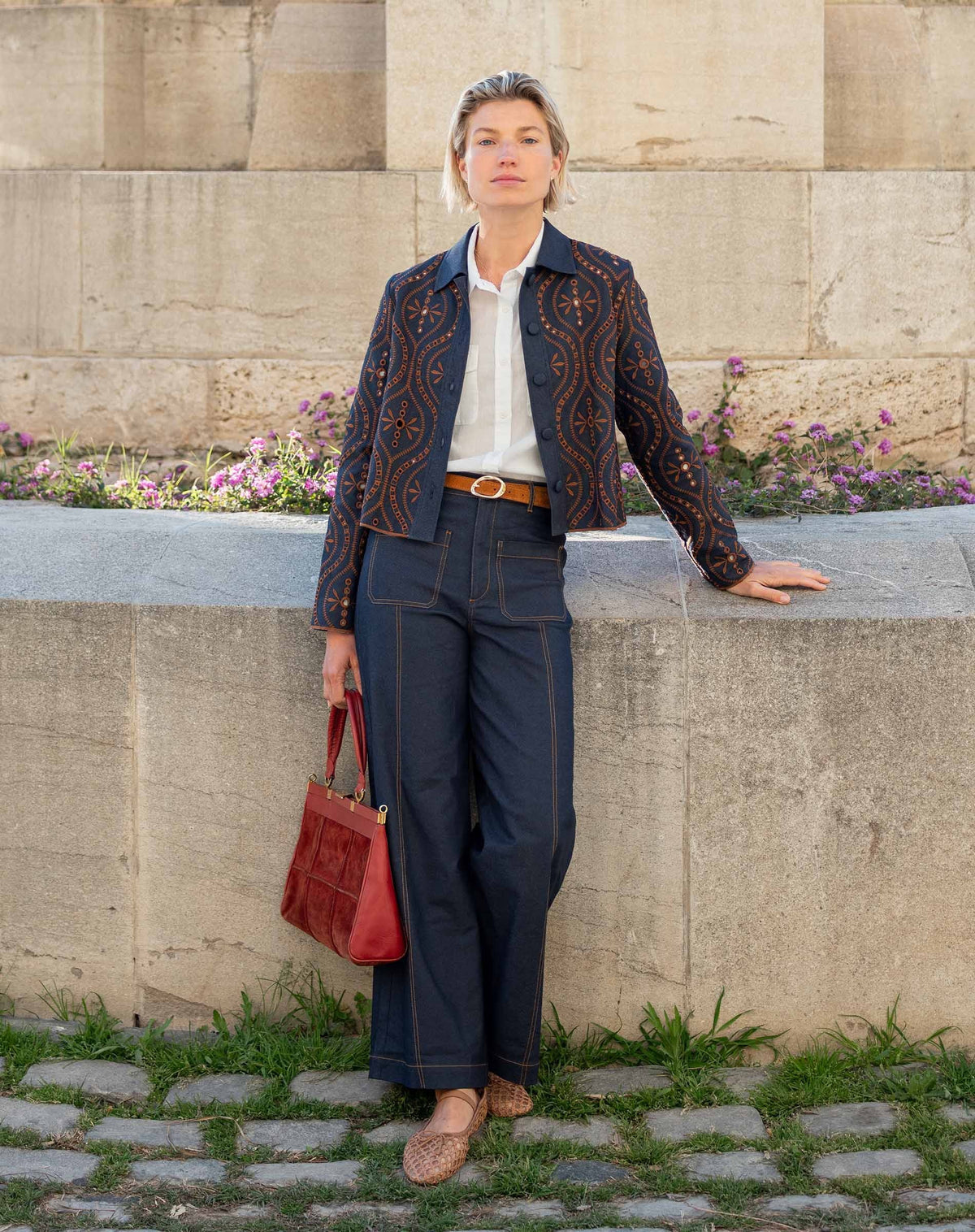 A woman with curly hair stands smiling in a bright room, wearing the MERSEA Andie Woven Pant in Rinse—high-waisted, wide-leg trousers with front pockets—paired with a white button-up shirt tucked in, a brown belt, and brown woven flats. One hand rests in her pocket.