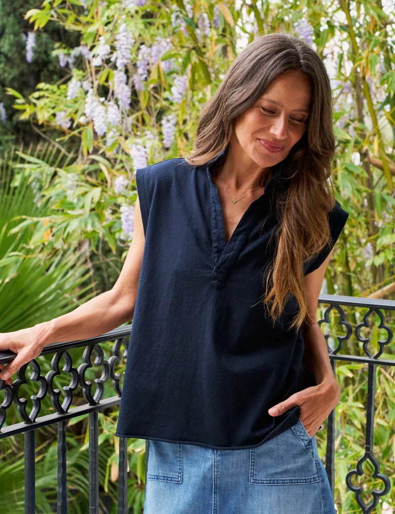 A woman with long brown hair stands on a balcony, smiling, wearing the FRANK & EILEEN Olivia Popover Henley Tank in British Royal Navy, with green foliage and purple flowers in the background.