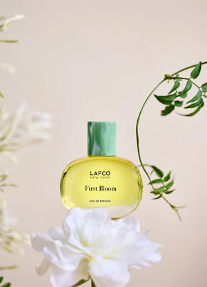 A box of LAFCO - FINE FRAGRANCE, FIRST BLOOM 50ML stands among white flowers and green vines against a pale beige background.