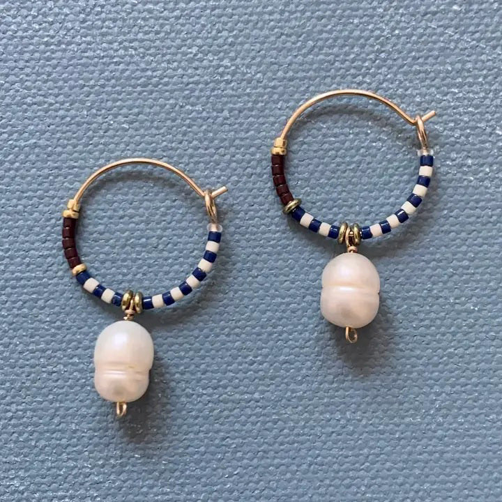 The ALICE RISE - PEARL DROP HOOPS by ALICE RISE are 14k gold filled hoops with blue, white, and brown Japanese glass beads and a central white pearl charm, beautifully displayed on a textured light blue surface.