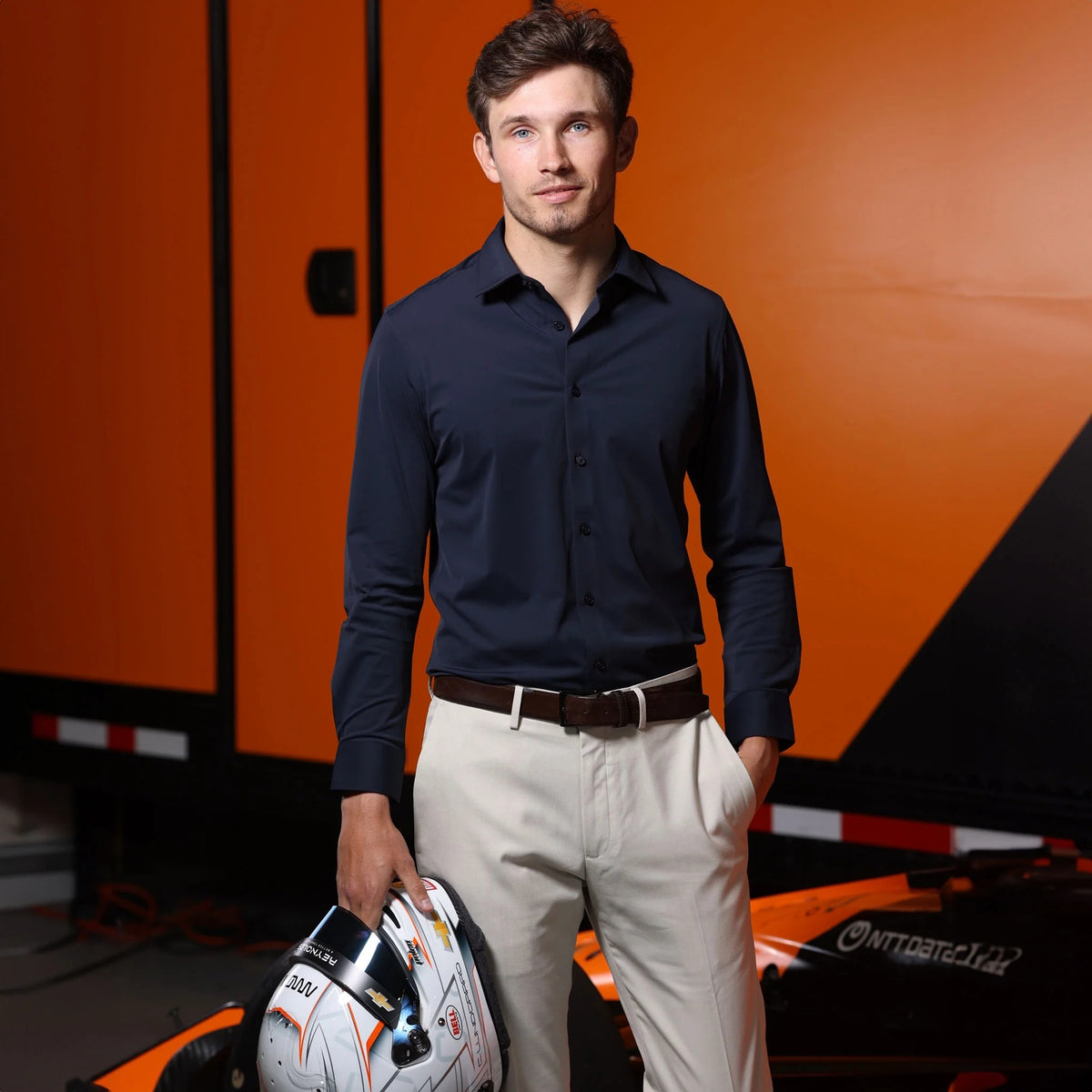 A man wearing the COLLARS AND CO - APEX PERFORMANCE DRESS SHIRT SEMI SPREAD IN NAVY stands in front of an orange race car holding a racing helmet, with an orange and black background behind him.