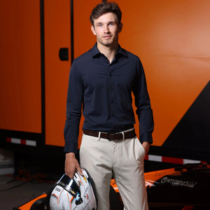 A man wearing the COLLARS AND CO - APEX PERFORMANCE DRESS SHIRT SEMI SPREAD IN NAVY stands in front of an orange race car holding a racing helmet, with an orange and black background behind him.