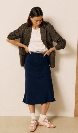 A woman with long brown hair and a neutral expression stands against a two-tone wall, wearing a grey sweater layered over a white shirt, white socks, pink shoes, and the HARTFORD - JIMA Bias Cut Skirt in Night by HARTFORD, effortlessly showcasing chic style with this navy midi skirt.