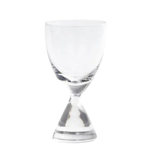 The ZODAX TIMELESS SIP SHOT GLASS, featuring a unique double-ended shape, sits half-filled with amber liquid on a light surface, while two dark round dishes are softly blurred in the background.