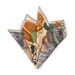 A woman in a beige short-sleeved top and matching skirt stands against a light background, adorned with the SABINA SAVAGE ORANGES AND LEMONS SILK TWILL SCARF in Cintrine/Zest. Her shoulder-length brown hair softly frames her face, enhancing her effortless elegance.