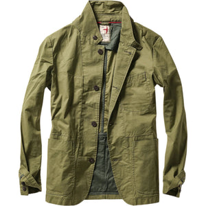 A person models the RELWEN Flyweight Flex Blazer in Burnt Olive over a blue and white checkered shirt with gray pants, hands in pockets, against a plain white background.