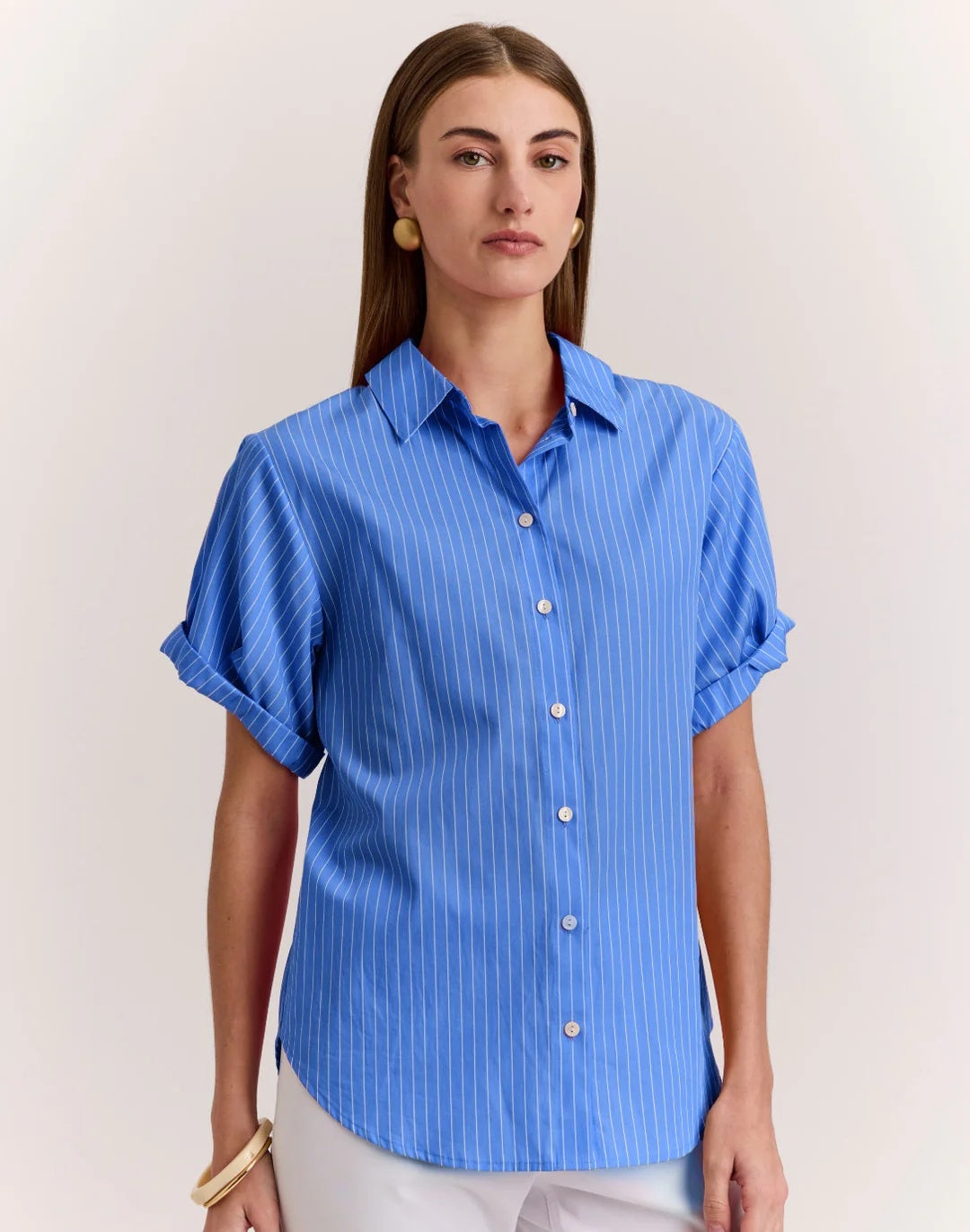 A woman wears the HINSON WU Lucie Short Sleeve Shirt in Pencil Stripe Lake Blue, crafted from 100% cotton, paired with white pants. She has straight brown hair and gold earrings, standing against a plain background with a neutral expression.