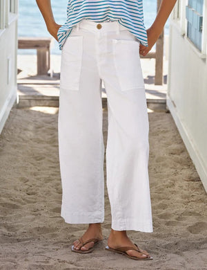 A person wearing the FRANK & EILEEN Limerick Dropped Wide Leg Carpenter Pant in Italian Performance Linen White, paired with a blue and white striped top and flip-flops, stands on sandy ground between two white walls with the ocean visible in the background. The cropped length of these FRANK & EILEEN pants enhances the breezy, relaxed feel of the seaside scene.