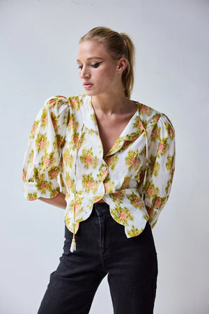 A woman with blonde hair in a ponytail wears the HOPE FOR FLOWERS Scalloped Shirt in Pink Warped Ikat by HOPE FOR FLOWERS and black high-waisted jeans, standing against a plain light background with her hands on her hips.