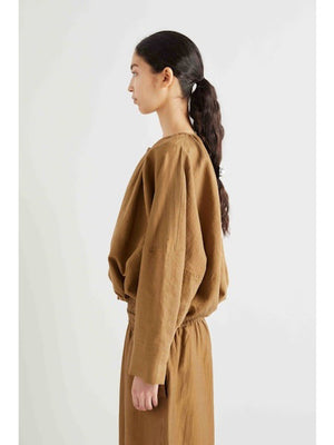 A person with long, dark hair in a low ponytail, held by a silver clip, is seen from behind wearing the MEIMEIJ - COTTON BOMBER JACKET in brown. The background is plain white.