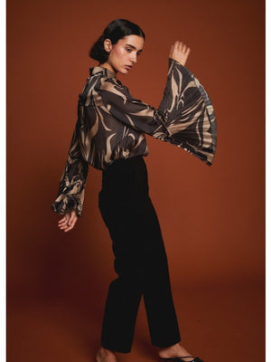 A woman poses against a brown background wearing the MILVA MI black and gold flare sleeve blouse by MILVA, paired with high-waisted black pants. Her dark hair is styled in a low bun.