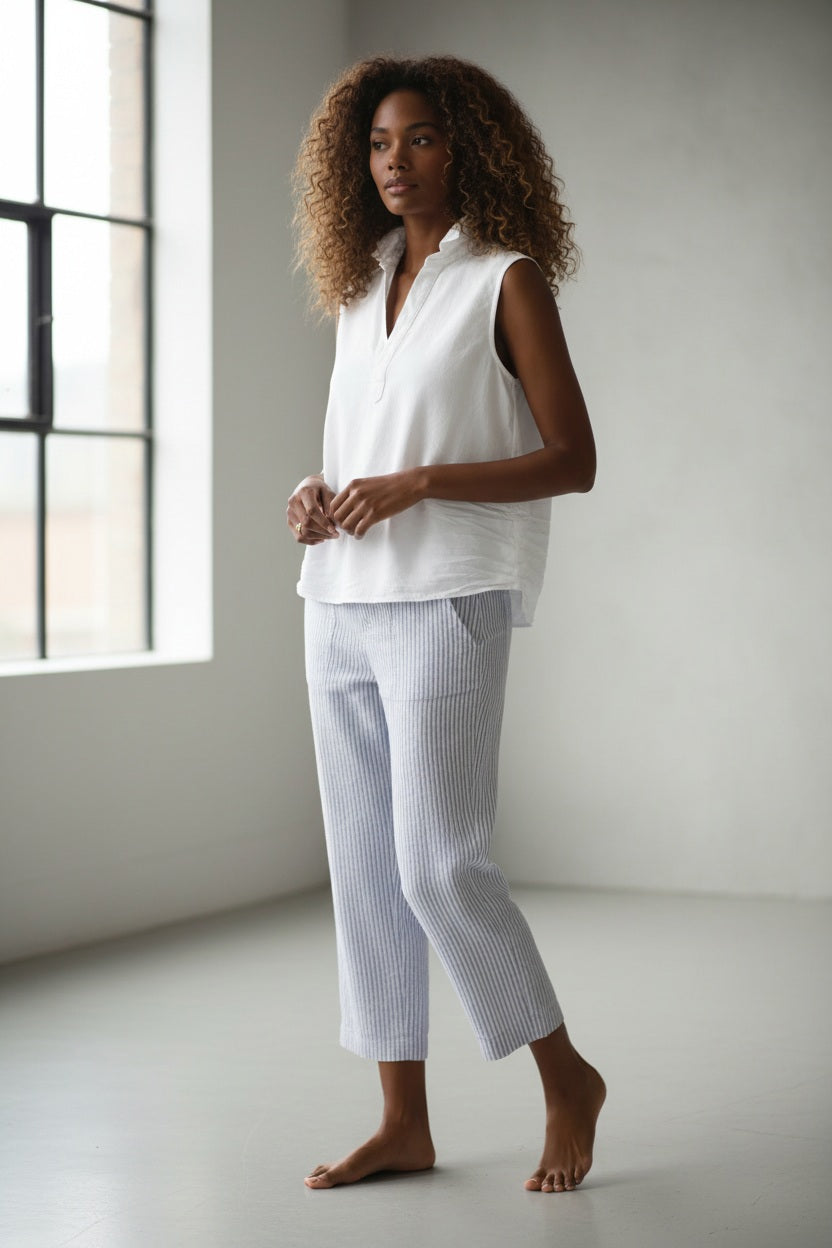 A woman with curly hair stands outdoors in front of green foliage wearing the FRANK &amp; EILEEN Olivia Popover Henley Tank in Famous Denim White, paired with a light-colored striped skirt.