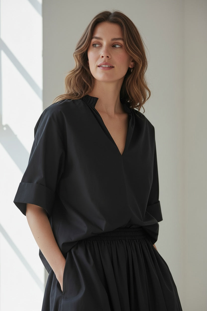 A woman with her hair in a bun stands in profile, wearing the MEIMEIJ Gathered Neck Blouse in Black by MEIMEIJ against a plain light background.