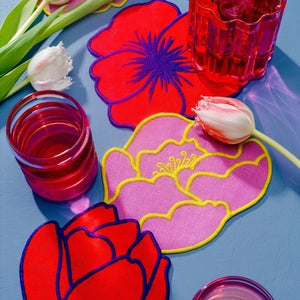 A box of "LINEN FLOWER COCKTAIL NAPKINS" by PIECEWORK PUZZLES features 100% linen napkins delicately embroidered with floral designs, surrounded by vibrant blossoms and colorful glassware on a light blue surface, creating a cheerful summer vibe.