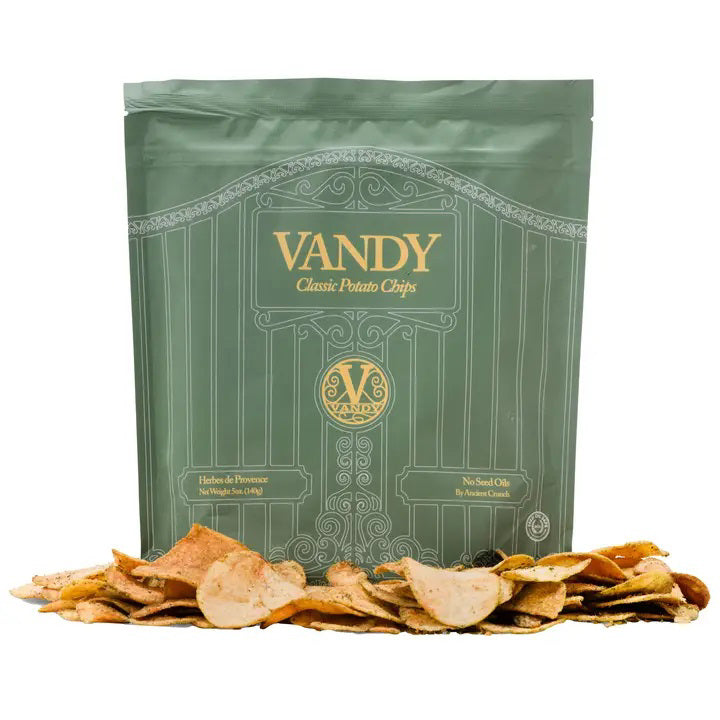An upright green bag of ANCIENT CRUNCH - VANDY POTATO CHIPS, HERBES DE PROVENCE features gold accents and a gate design, with tallow-cooked potato chips scattered at its base.