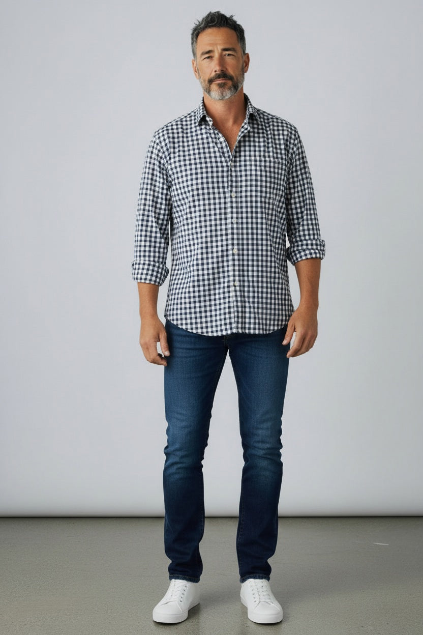 A man with short dark hair and a beard wears the FRANK &amp; EILEEN Finbar Men’s Button Up Shirt in Italian Chambray Blue Check, paired with blue jeans and white sneakers, standing against a plain light gray background.